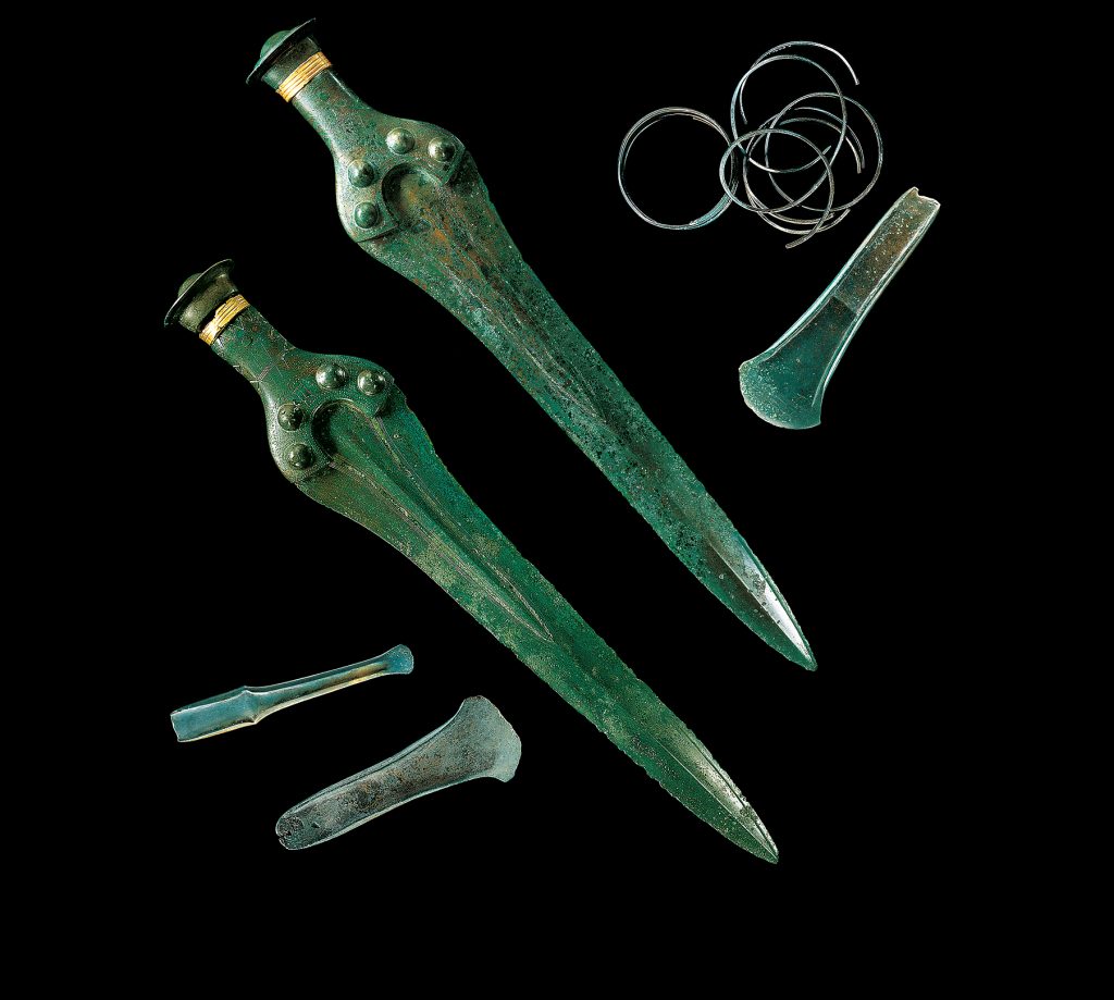 Zwei dolchartige Schwerter, drei Beilköpfe und eine Sammlung von dünnen Armreifen aus Bronze. Das Material schimmert grünlich. Die Gegenstände wurden zusammen mit der Himmelsscheibe in Nebra ausgegraben und sind mindestens 3600 Jahre alt.