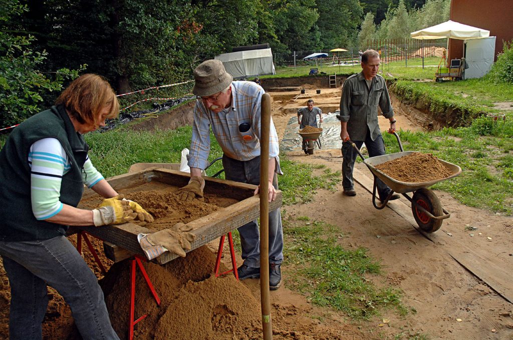 Ausgrabungen in Kalkriese dem historischen Ort der Varusschlacht. Zwei Männer mit Schubkarren voller Erden und zwei Personen im Vordergrund, die Erde nach Fundstücken sieben.