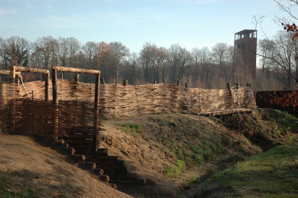 Bild aus dem Landschaftspark des Museums Kalkriese, dem Ort der Varusschlacht. Man sieht einen Zaun aus geflochtenem Holz und Erdwälle. Im Hintergrund der Turm des Museum.