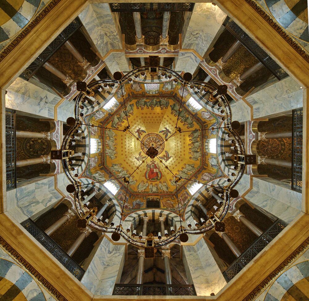 Von unten nach ober blickend hat der Fotograf im achteckigen Innenraum des Aachener Doms die reich verzierte Kuppel und den Leuchter fotografiert.