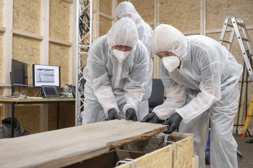 Zwei Wissenschaftler in Schutzanzügen und mit Mundschutz heben im Juni 2025 den Deckel des mittelalterlichen Holzsarges von Otto dem Großen ab.