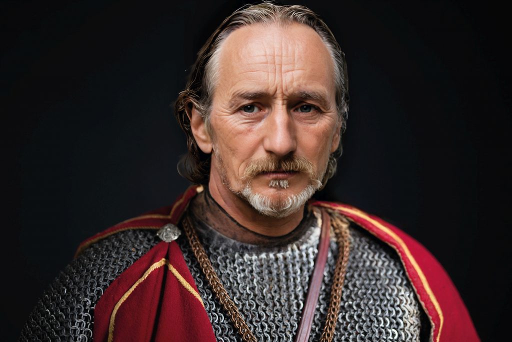 Ein Portraitfoto eines älteren Mannes mit graumelierten Haaren und einem gestutzten Bart. Er trägt ein Kettenhemd mit einem roten Umhang darüber. Das Bild ist eine KI Nachbildung von Heinrich II. So soll er ausgesehen haben.