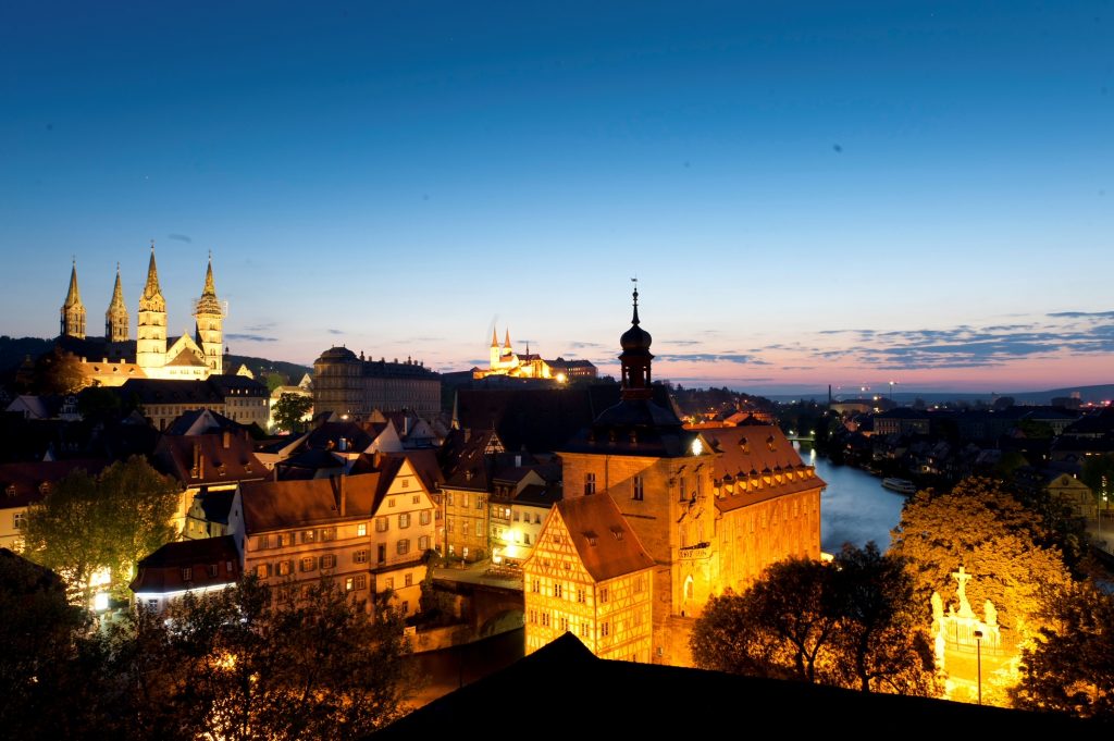 Die mittelalterliche Stadt Bamberg bei Nacht, von oben fotografiert. Man sieht Fachwerkhäuser, die Regnitz und den angeleuchteten Bamberger Dom