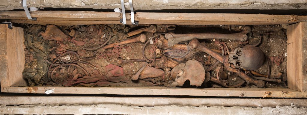 Blick in den geöffneten Holzsarg von Otto dem Großen, dem ersten deutsch-römischen Kaiser. Man sieht ein aufgelöstes Skelett mit einem gut erhaltenen Schädel. Zusätzlich sind Reste von Textilien zu sehen.